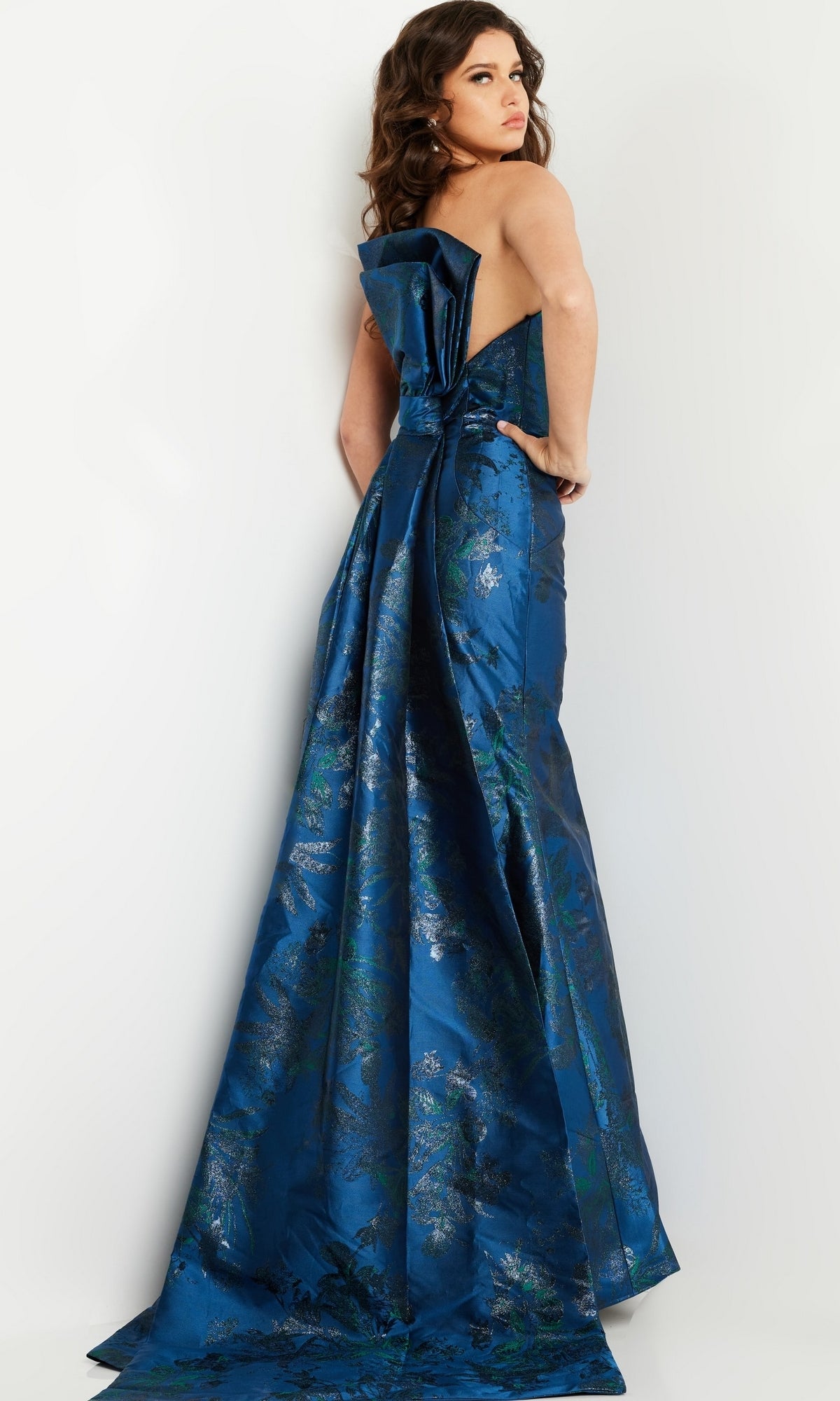 A woman models the Jovani 23898 Navy Blue Long Formal Dress, featuring a strapless design, large bow, and flowing train, as she stands against a light background looking over her shoulder.